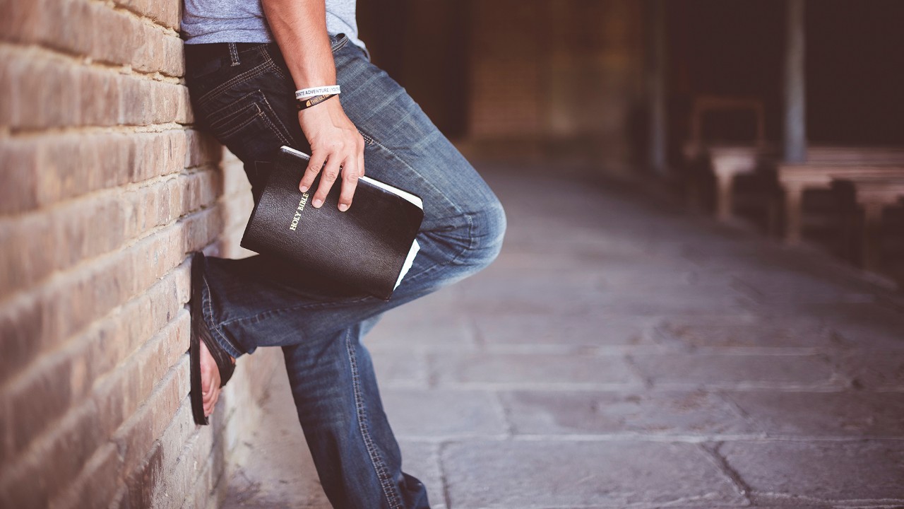 Man holding bible leaning against a brick wall.